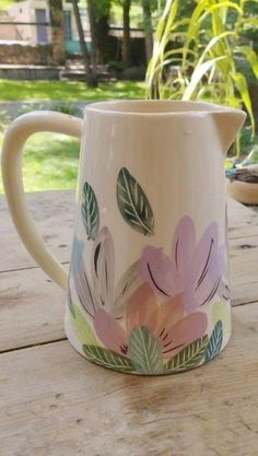 a white pitcher with pink flowers painted on it sitting on a wooden table in front of some plants