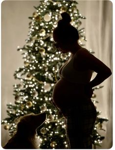 a pregnant woman standing in front of a christmas tree with her dog looking at her belly