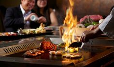 a man and woman cooking food on a grill with flames in the air behind them