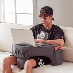 a young man sitting on a couch using a laptop computer while wearing a baseball cap