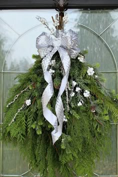 a wreath with white flowers and silver ribbons hanging on a glass front door, decorated with greenery