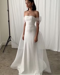 a woman standing in front of a white backdrop wearing a wedding dress with off the shoulder sleeves