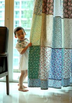 a little boy standing in front of a window next to a curtain covered with colorful fabric