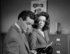 a man and woman looking at something on a desk in an office setting, one holding a clipboard while the other looks down