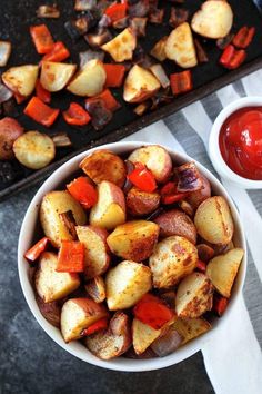 a white bowl filled with cooked potatoes next to a tray of ketchup on a table