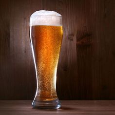 a tall glass filled with beer sitting on top of a wooden table next to a wall