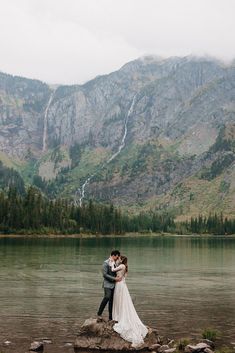 a bride and groom standing on rocks in front of a lake with mountains behind them