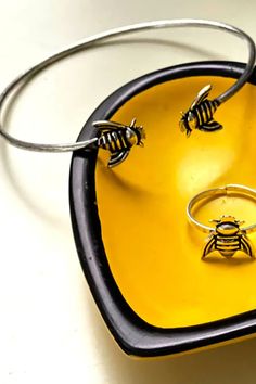 two bee rings sitting on top of a yellow plate next to a pair of silver bracelets