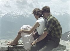 a man and woman sitting on top of a rocky mountain with snow capped mountains in the background