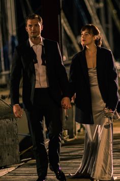 a man and woman walking down a street at night with boats in the background,