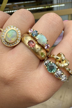 Close-up of a hand wearing five yellow-gold rings, each with a different style and type of gemstone. Two have a row of four claw-set stones, one is a 'pretzel' knot ring studded with white diamonds, one is a wide band with inset gemstones and  the last has a large central oval opal with a white diamond halo. Mothers Day Jewelry Gifts, Ring Inspo Jewelry, Family Heirloom Jewelry, One Piece Jewelry, Five Gold Rings, Gold And Light Blue, Wedding Stack, Cool Rings, Famous Jewelry