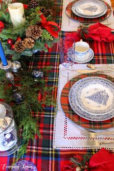 the table is set for christmas dinner with red and green plaid placemats, silverware, pine cones, candles, and evergreen branches