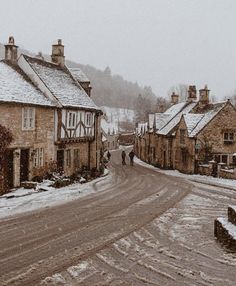 two people walking down a snowy street in the middle of some small village buildings with snow on them