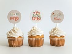 three cupcakes with white frosting on top and one topped with a sticker