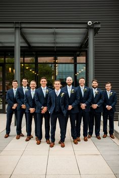 a group of men standing next to each other in front of a black building and wearing suits