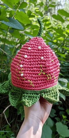 a hand is holding up a crocheted hat in front of some leaves and bushes