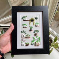 a person holding up a framed artwork with rocks and plants on it in front of a window