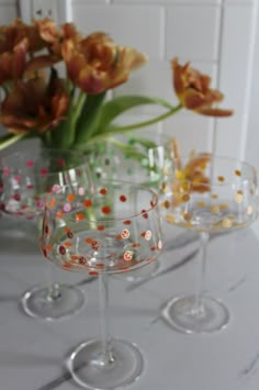 three wine glasses sitting on top of a table with flowers in the vase behind them