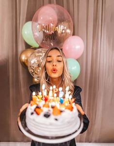 a woman holding a cake with candles in front of her face and balloons behind her