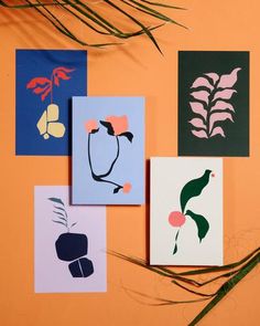 four cards with different designs on them sitting next to some plant stems and leaves in front of an orange wall