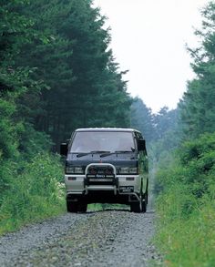 a van is driving down a gravel road in the middle of tall grass and trees