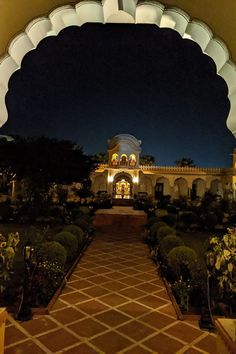 an archway leading to a building with many plants and flowers on the ground at night