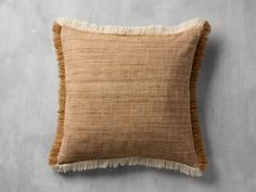 a tan pillow with fringe on it against a gray wall, close up view from the front