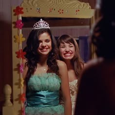 two young women standing in front of a mirror wearing dresses and tiaras, smiling at the camera