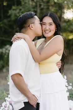 a man and woman standing next to each other in front of some flowers with their arms around each other