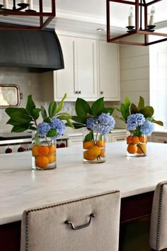 three vases filled with flowers and oranges on a kitchen counter