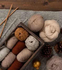 several balls of yarn in a wooden box next to some pine cones and knitting needles