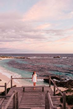 a woman standing at the end of a wooden walkway next to the ocean on a cloudy day