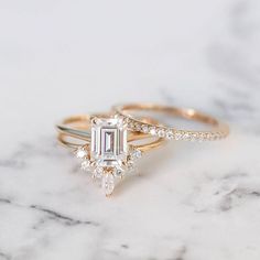 two gold engagement rings with an emerald cut diamond on the top and side, sitting on a marble surface