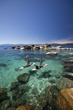 two people are standing on surfboards in the clear blue water near rocks and boulders