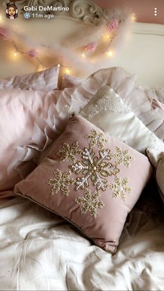 a bed topped with lots of pillows covered in pink and white blankets next to a string of lights