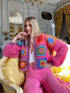 a woman is sitting on a couch wearing pink pants and a colorful cardigan sweater