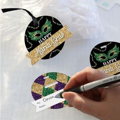 a hand holding a marker next to two mardi gras tags with the words happy mardi gras on them