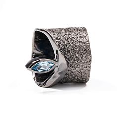 A dazzling blue topaz pierces the top of this black rhodium plated silver ring. It is ringed again in rhodium and emerges from a band that encircles the ring. The barrel is undulating and textured with natural shape and form which echoes true nature. Metal: 925 Silver Stones: Blue Topaz Plating: Black Rhodium  PLEASE NOTE: This piece is handmade and may have slight variations from the displayed image. Natural gemstones can differ in shape and color depending on lighting. For an accurate represen Nature Details, August Birthstone Jewelry, July Birthstone Jewelry, Fine Art Jewelry, Pearl Jewellery Earrings, Jewelry Ring Box, Men's Jewelry Rings, True Nature, Black Rhodium