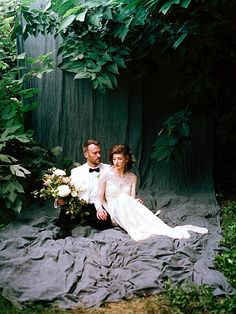 a man and woman are sitting on a blanket in the grass with greenery behind them