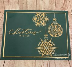 a green and gold christmas card with ornaments hanging from it's sides on a wooden surface