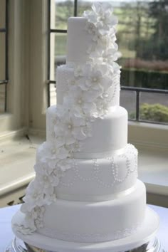 a large white wedding cake with flowers on the top and bottom tier is sitting in front of a window