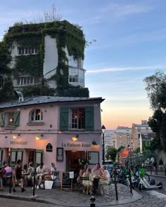 people are sitting outside on the sidewalk in front of a building with plants growing up it's side