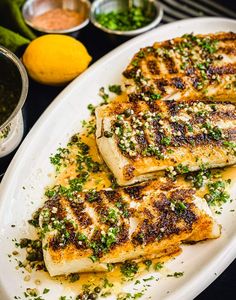 grilled fish fillets on a white platter with lemons and seasonings