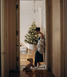 a woman standing next to a baby in front of a christmas tree with a teddy bear on the floor