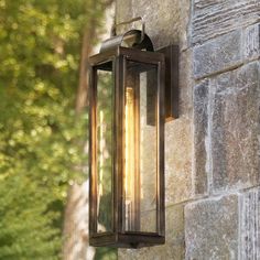 an outdoor light on the side of a stone building with trees in the background and sunlight shining through it