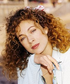 a close up of a person with curly hair wearing a blue shirt and jeans leaning on a wall