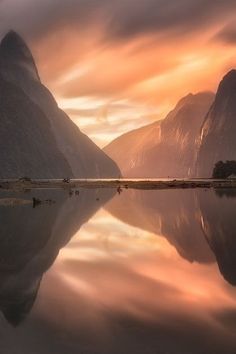 the mountains are reflected in the still water at sunset, with one person walking on the shore