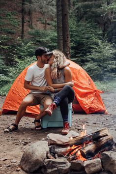 a man and woman sitting next to a campfire in front of an orange tent