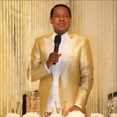 a man in a gold suit holding a microphone at a table with white flowers and candles