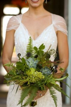 a woman in a wedding dress holding a bouquet of flowers and greenery on her arm